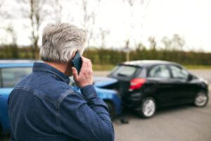 A man on the phone, looking at his smashed car, wondering why he needs a car accident lawyer.